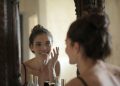 Woman smiling while touching her face in the mirror, enjoying a skincare routine.