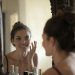 Woman smiling while touching her face in the mirror, enjoying a skincare routine.